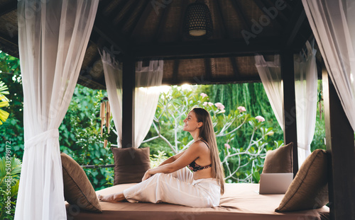 Side view of cheerful female digital nomad dressed in stylish apparel smiling during leisure pastime at patio terrace, carefree Caucasian freelancer in casual clothes resting at courtyard