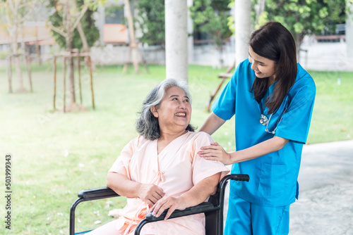 Asian doctor woman, attractive and young, looking and taking care elderly woman patient with paralysis, is sitting on a wheelchair, to elderly patient and health care concept.