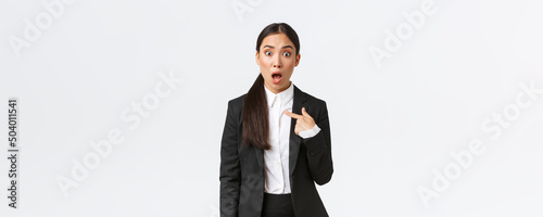 Shocked and offended young asian woman in suit pointing at herself with astounded panic face, being accused. Businesswoman looking insulted after being named, white background