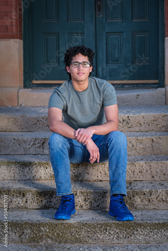 A young man sits in front of an old building with cement steps and a green door.