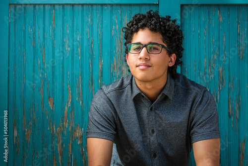 A young man with curly hair sits in front of a weathered blue wood wall.