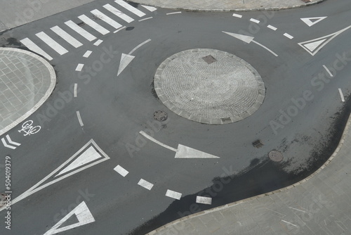 View from top on empty traffic circle or roundabout road with white arrows, crosswalk and other road signaling or marking in European town. Grey asphalt is partly wet after rain.