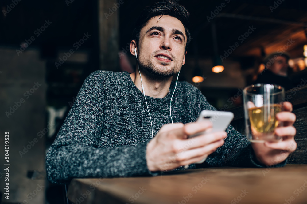 Contemplative hipster guy in digital earphones thinking about audio content during weekend leisure in cafeteria, millennial male blogger with beverage and mobile gadget thoughtful looking away
