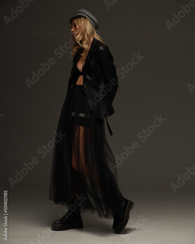 Fashionable american blonde pretty woman in black eco leather boots, jacket, transparent tulle skirt, studio shoot