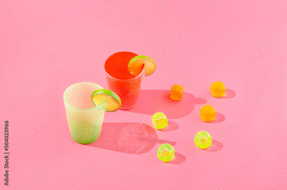 A lime green and a tangerine orange glass with lemon slices and colorful ice cubes against warm pink background. Moder creative concept for summer mood banner or wallpaper A lime green and a tangerine orange glass with lemon slices and colorful ice cubes against warm pink background. Moder creative concept for summer mood banner or wallpaper