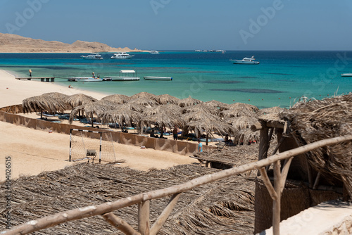 Holidays in Egypt. Beach with umbrellas made of dry palm leaves, desert and yachts in the blue water of the Red Sea. Vacation and Holidays in Egypt. © PhotoRK