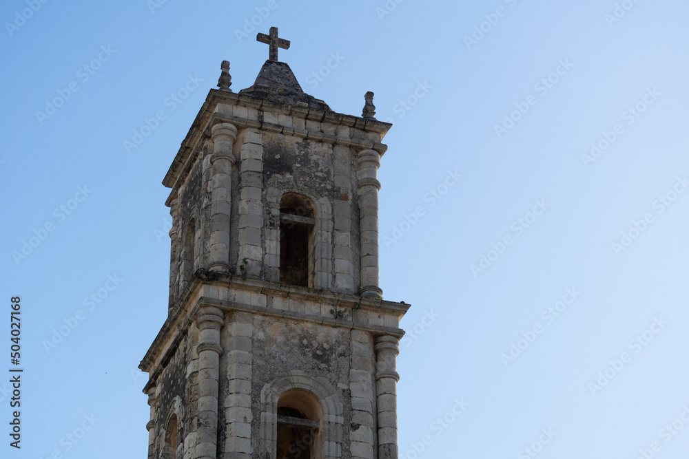 The tower of the old catholic church built in the colonial style. Bell tower with a crucifix on the roof against the blue sky.