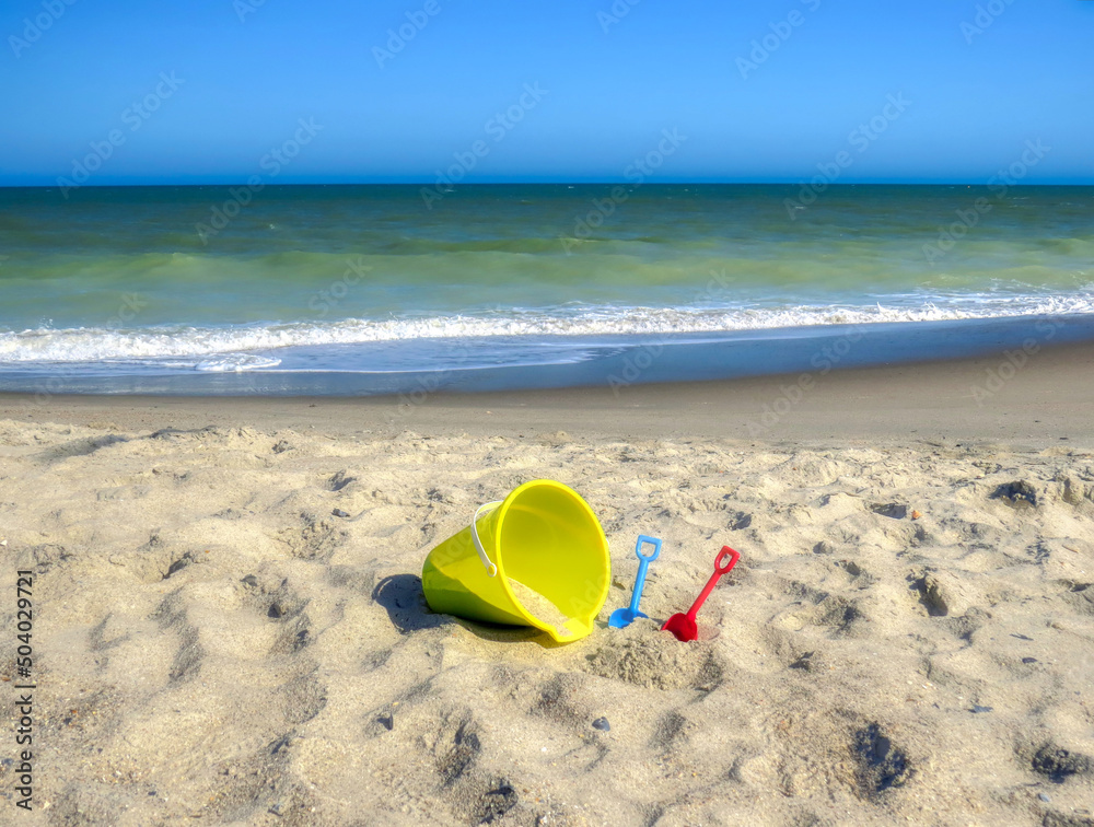 A yellow sand bucket and colorful toy shovels at the beach by the ocean. A yellow sand bucket and colorful toy shovels at the beach by the ocean.