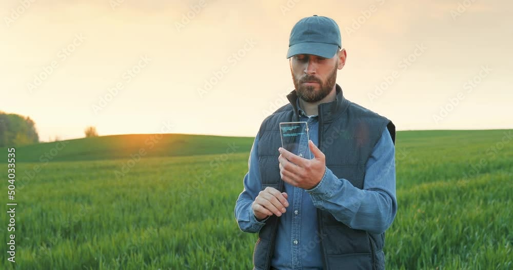 Confident caucasian man in casual attire and cap standing among green field, holding glass futuristic smartphone with holograms and tapping on screen. Agronomy and innovative technology concept. Confident caucasian man in casual attire and cap standing among green field, holding glass futuristic smartphone with holograms and tapping on screen. Agronomy and innovative technology concept.