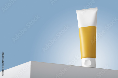 Care cream tube mockup on white table or podium, blue backdrop. Blank yellow label. Sunscreen treatment cosmetics template, copy space