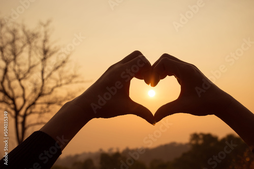 A couple hand making heart shape with the background of sky and the sun.