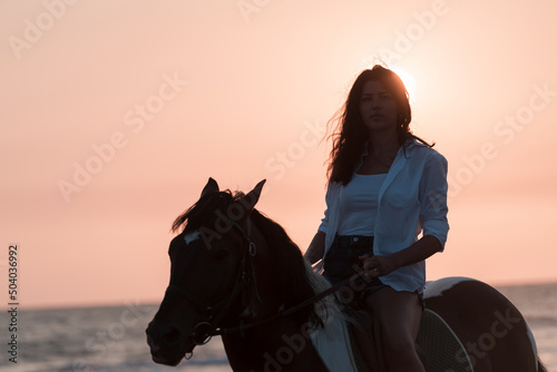 Woman in summer clothes enjoys riding a horse on a beautiful sandy beach at sunset. Selective focus 