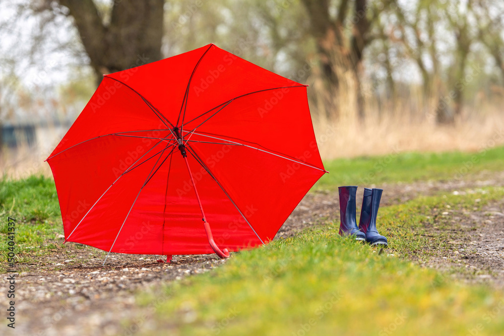 Bad weather and rainy days: Focus on blue wellies or rubber boots and a red umbrella on a rural dirt road in early spring outdoors