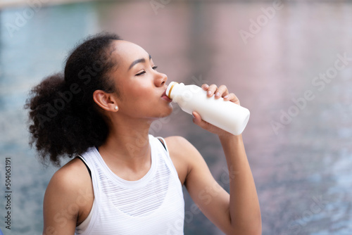 Young black woman with curly hair drinking fresh milk while sitting beside the swimming pool after workout for burning. Need protein, calcium, and good fat to build her muscles and control food, sugar