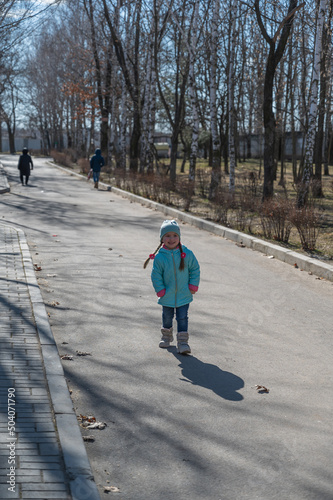 A child having fun in a sunny spring park. A five-year-old girl with long pigtails in a turquoise jacket poses and curses. A birch tree without leaves in the background