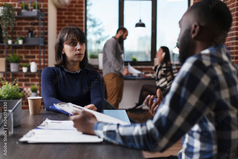 Business company recruiter and candidate sitting at desk in office while reviewing job applicant CV. Multiethnic people in startup agency talking about salary offer and internal regulations. Business company recruiter and candidate sitting at desk in office while reviewing job applicant CV. Multiethnic people in startup agency talking about salary offer and internal regulations.