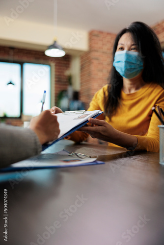 Businesswoman with medical face mask holding clipboard while freelancer signing papers for executive manager job during company interview. Team working at business collboration in startup office photo