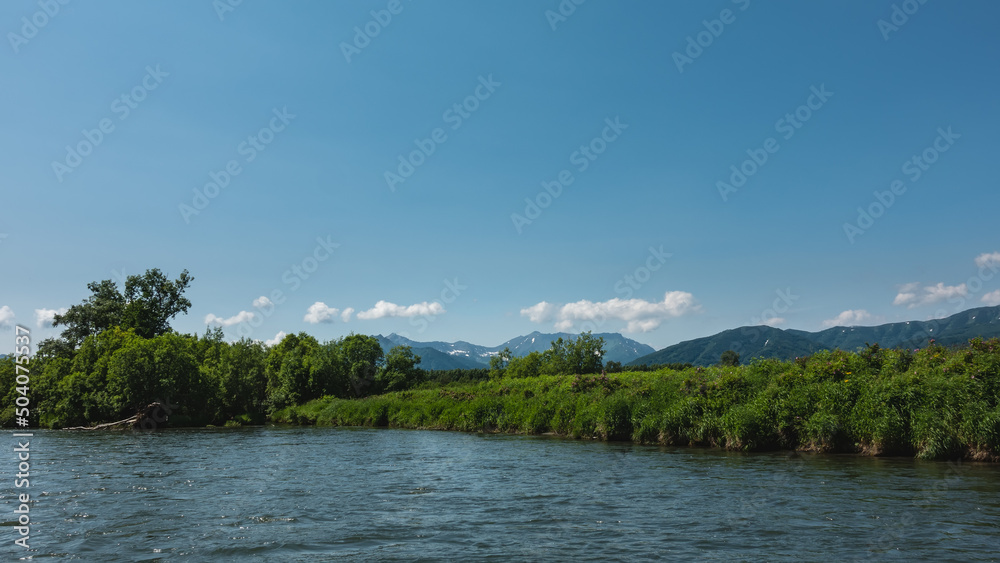 Blue calm river. Lush grass and green trees grow on the shore. In the distance, against the background of the azure sky and clouds, a mountain range is visible. Kamchatka Blue calm river. Lush grass and green trees grow on the shore. In the distance, against the background of the azure sky and clouds, a mountain range is visible. Kamchatka