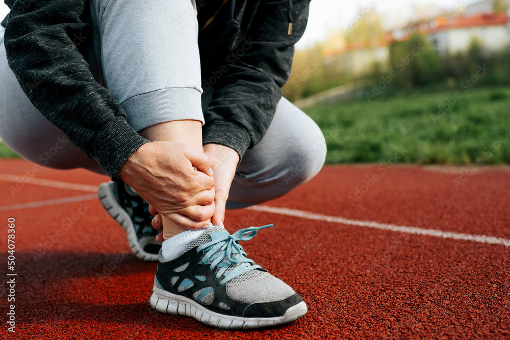 Woman runner holding leg suffering from muscle and tendon sprain pain in ankle at outdoor stadium, close-up. Athlete injured during intense workout. Health care and sport concept - obrazy, fototapety, plakaty 