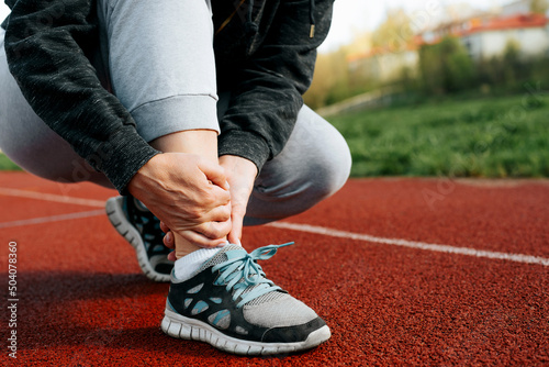 Woman runner holding leg suffering from muscle and tendon sprain pain in ankle at outdoor stadium, close-up. Athlete injured during intense workout. Health care and sport concept photo
