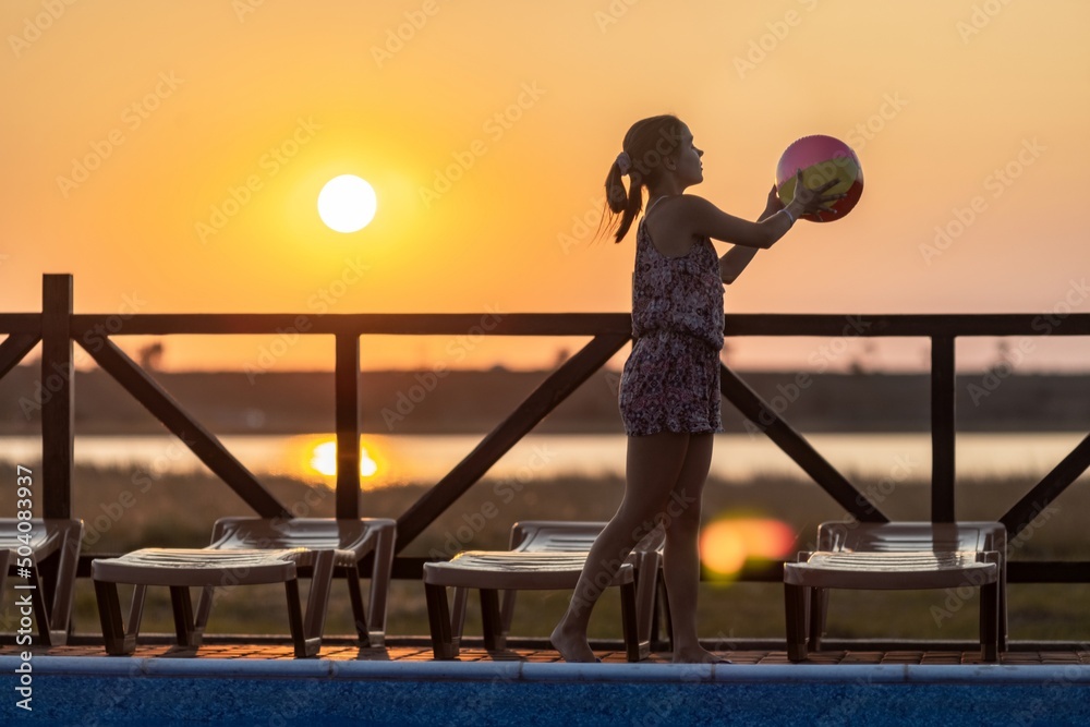 Fototapeta premium A girl with hair braided in a bun in bright suit plays by the pool with a ball against the background of summer sun