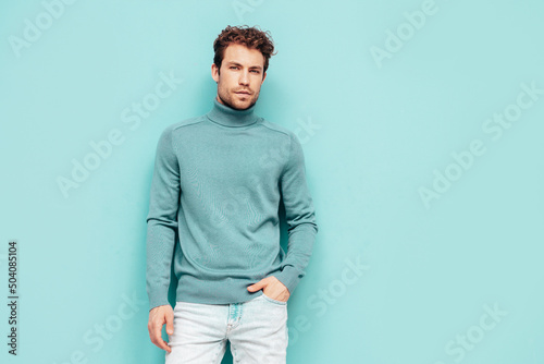 Portrait of handsome confident  model. Sexy stylish man dressed in  sweater and jeans. Fashion hipster male with curly hairstyle posing near blue wall in studio. Isolated