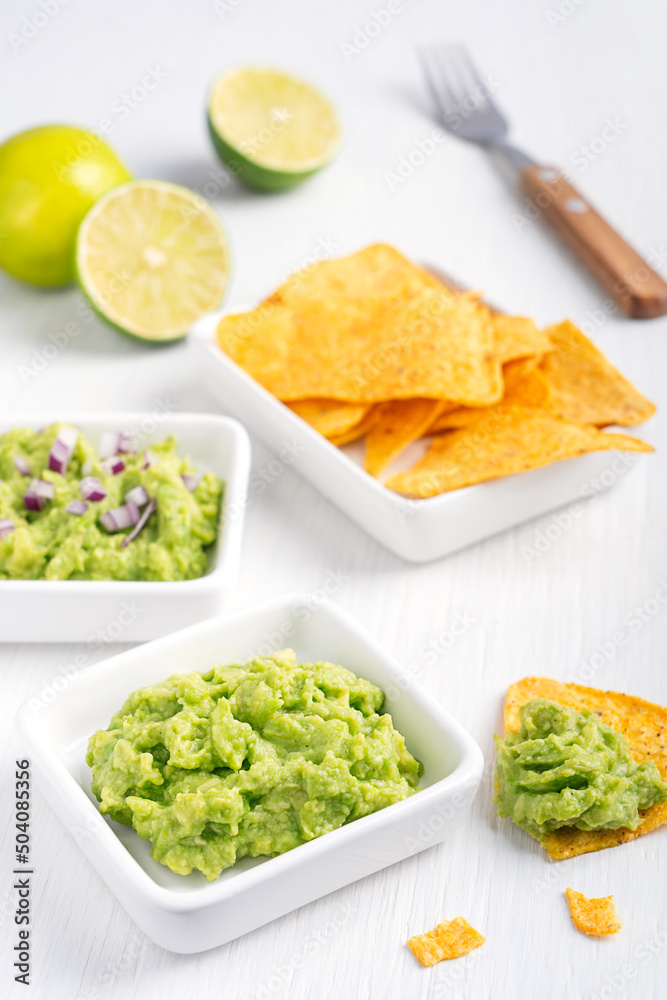 Two types of vegan mexican guacamole dip, sauce or salad made of pureed avocado with red onion served with nachos or tortilla chips on white wooden table with fork and lime citrus as healthy snack