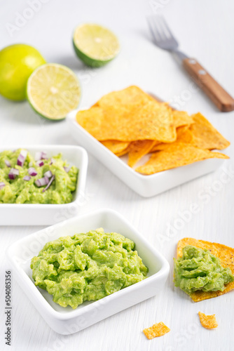 Two types of vegan mexican guacamole dip, sauce or salad made of pureed avocado with red onion served with nachos or tortilla chips on white wooden table with fork and lime citrus as healthy snack