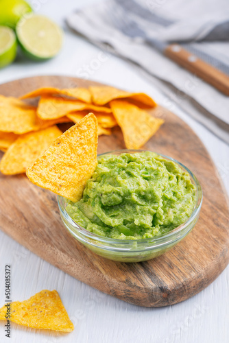 Homemade organic mexican guacamole dip, sauce or spread made of mashed ripe avocado served with crunchy nachos or tortilla chips on cutting board on white wooden table as snack with lime and towel