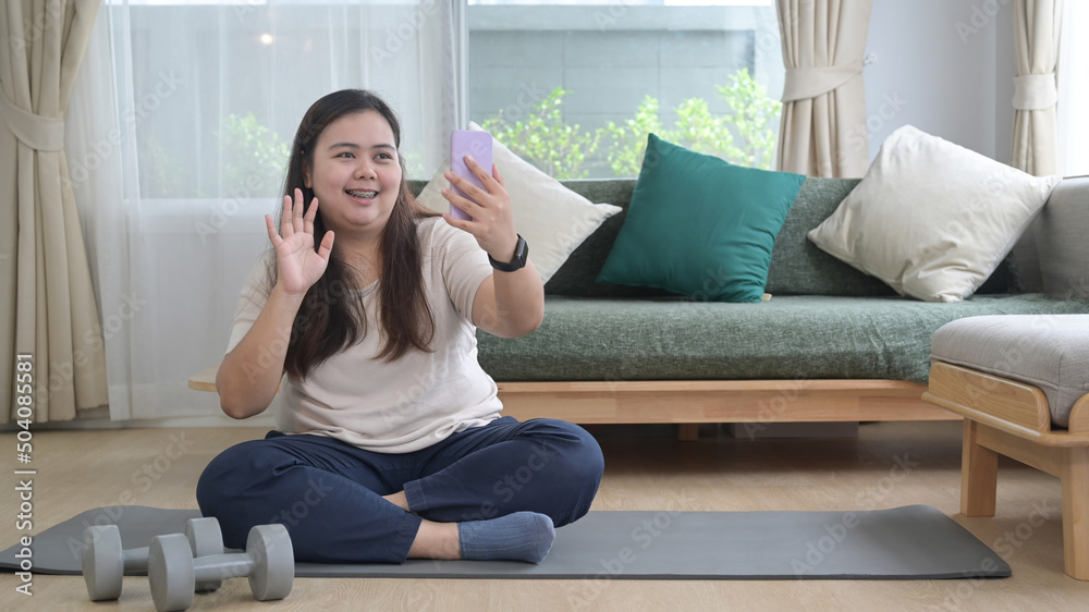 Cheerful overweight woman sitting on mat in living room and making video call via her smart phone. Cheerful overweight woman sitting on mat in living room and making video call via her smart phone.