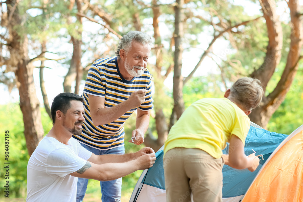 Senior man putting up camping tent with his son and little grandson in forest Senior man putting up camping tent with his son and little grandson in forest