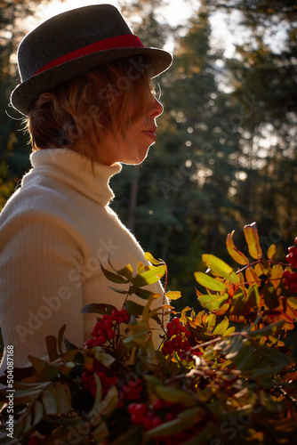 Cute girl in a white dress, black hat and with a bouquet of rowan branches and red berries in nature in the forest and light of sun on background. Photo shoot in nature landscape in sunny autumn day photo