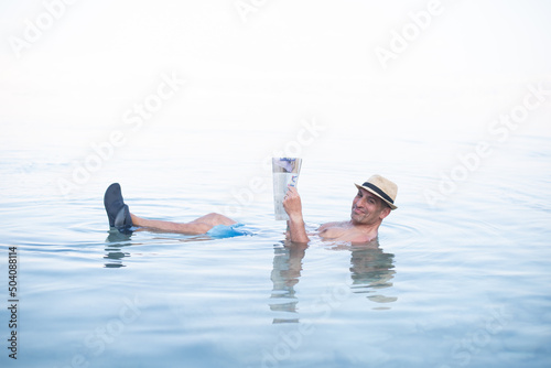 Man lies in blue water of Dead Sea. Person in hat and with newspaper. Very salty water push out. Treatment and recreation on beach, travel and holidays in Israel. Salt and mud healthy for body photo