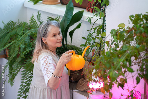 An elderly woman stands on a stepladder and waters indoor flowers from a watering can. A woman takes care of houseplants.