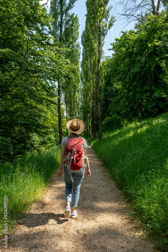 Woman with brown hair, gray t-shirt, jeans, straw hat and red backpack hiking in the nature in front of a avenue of trees, Staatspark Fürstenlager, Staatspark Fürstenlager, Auerbach, Bensheim, Germany © Bildgigant