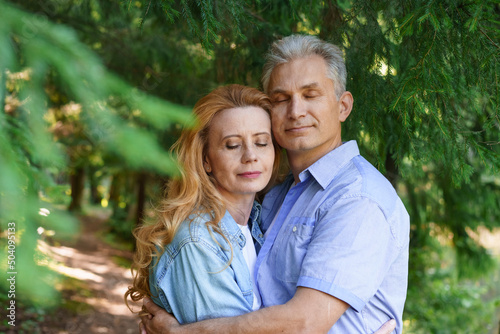 Smiling senior couple standing in park hugging each other together and smiling with happiness against the backdrop of trees on a warm day, enjoying retirement caucasian adult man and woman