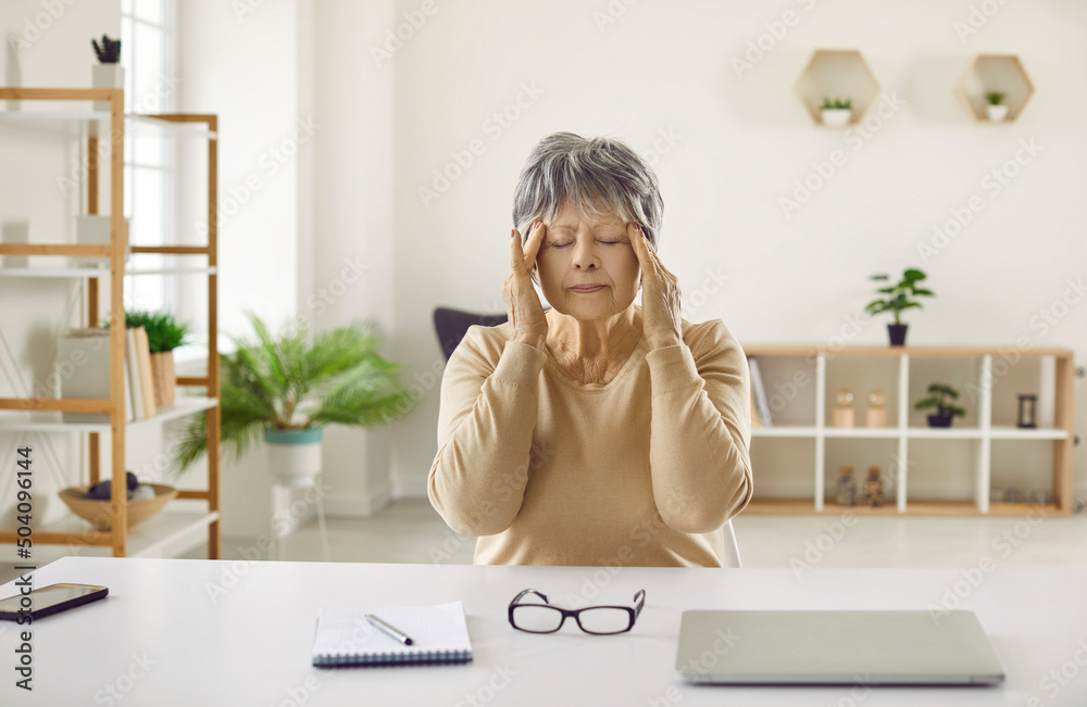 Tired senior woman sitting at table and doing self head massage. Exhausted mature lady who has headache after working on computer for a long time sitting with her eyes closed and rubbing her temples Tired senior woman sitting at table and doing self head massage. Exhausted mature lady who has headache after working on computer for a long time sitting with her eyes closed and rubbing her temples