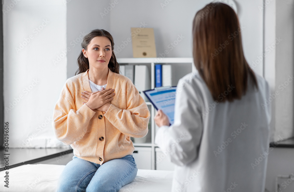 Fototapeta premium medicine, healthcare and people concept - female doctor with clipboard talking to woman holding to her chest patient at hospital