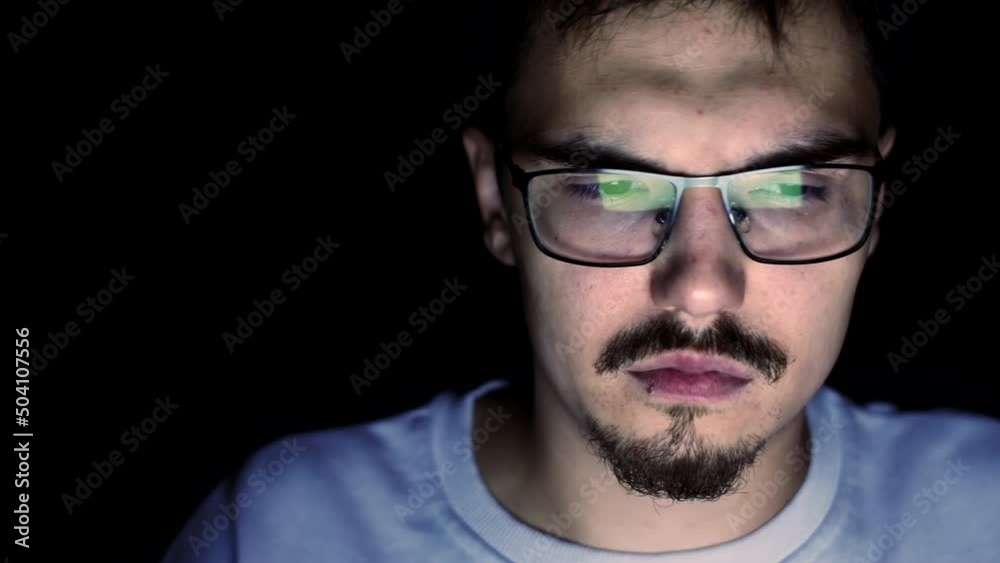 Young man with glasses is sitting at computer at night, tired and sad look, front view. Man with gambling addiction sits night on computer. Concept of dependency Young man with glasses is sitting at computer at night, tired and sad look, front view. Man with gambling addiction sits night on computer. Concept of dependency