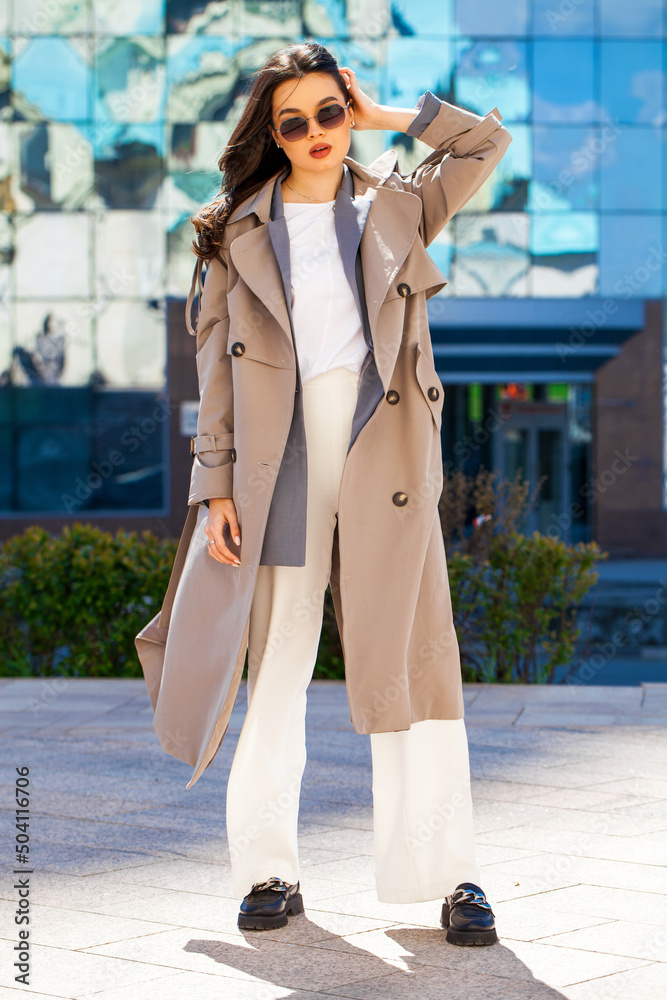 Portrait of a young beautiful brunette woman in gray coat and white trousers