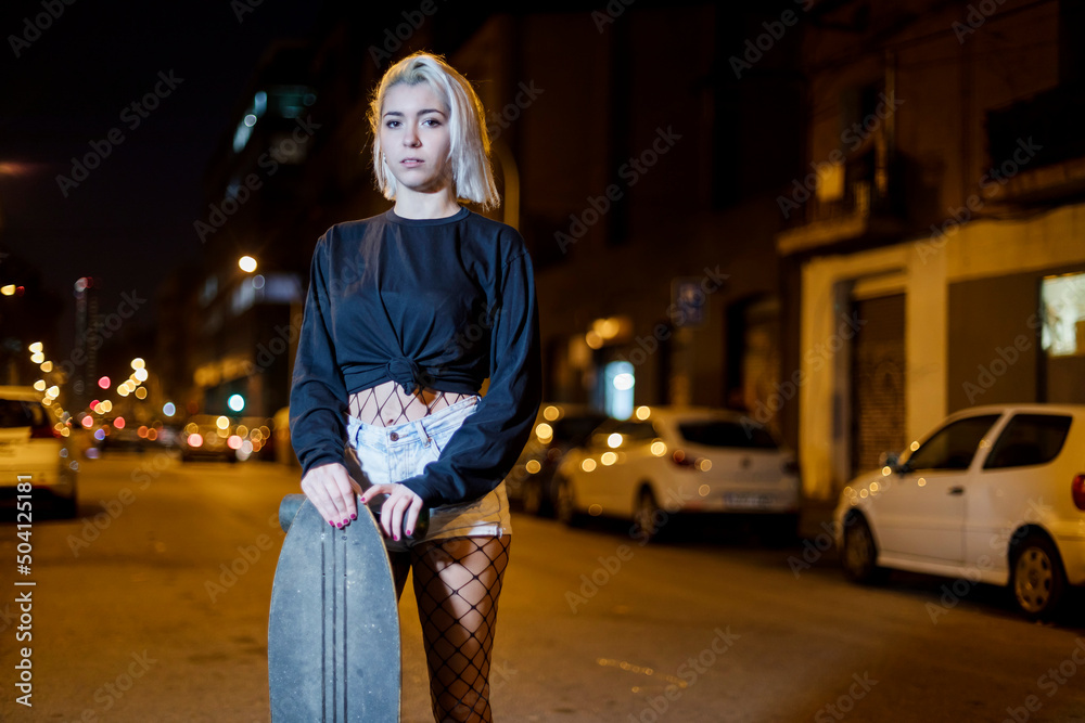 Fototapeta premium Front view of young pretty woman looking camera in shorts while standing on the street holding a skateboard at night in the city