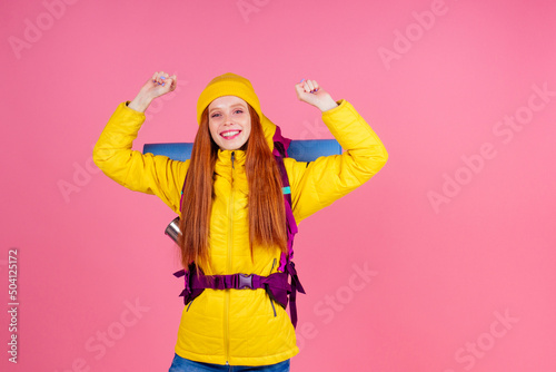 Female hiker with a backpack looking at the camera and smiling isolated on pink studio background.she wearing yellow windbreaker waterproof jacket and knitted hat © yurakrasil