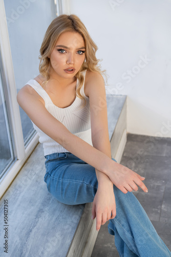 pretty woman in white tank top and jeans sitting on windowsill and looking at camera.