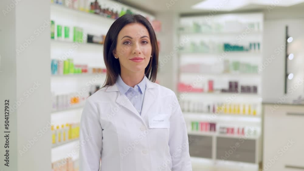 Beautiful caucasian woman in white lab coat turning head and looking at camera while standing among drugstore with medication on shelves. Concept of people, healthcare and pharmaceutics. Beautiful caucasian woman in white lab coat turning head and looking at camera while standing among drugstore with medication on shelves. Concept of people, healthcare and pharmaceutics.