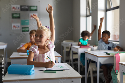 Multiracial elementary school students with hands raised sitting at desk in classroom
