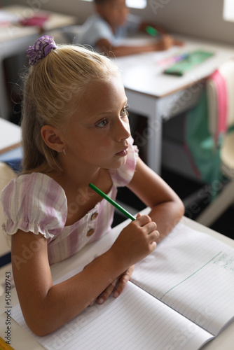 Close-up of caucasian elementary schoolgirl looking away while sitting with book and pencil at desk
