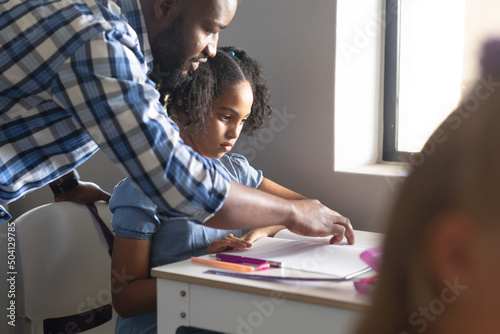 African american young male teacher teaching biracial elementary schoolgirl at desk in classroom