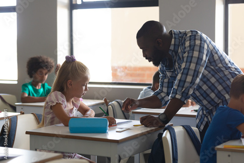 African american young male teacher teaching to caucasian elementary schoolgirl sitting at desk