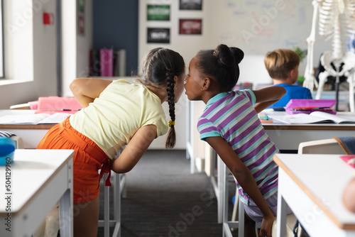 African american elementary girl whispering to caucasian female classmate at desk in classroom