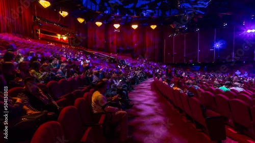 Spectators gather in the auditorium and watch the show in theatre timelapse. Large hall with red armchairs seats. Viewers filling places until turn off the light. Panoramic view from left side photo